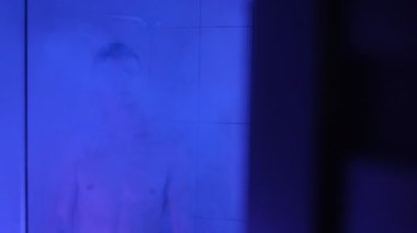 Duşta, duygusal olarak yorucu bir günün ardından, buhar ve soğuk mavi ışıkla kaplanmış, sorunlarını yansıtan bir adam..