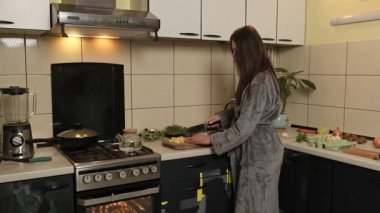 Yumuşak gri bornozlu bir kadın sıcak, ev yapımı bir mutfakta taze sebzeleri ahşap bir kesme tahtasıyla dikkatlice doğruyor ve ev yapımı yemek için malzemeler hazırlıyor..