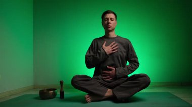 Nilüfer pozisyonundaki bir adam, elleri göğsünde ve karnında derin nefes almaya odaklanır. Renkli ışıklar değişirken rahatlama ve huzur yaşar ve meditasyon durumunu vurgular.