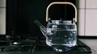 Gaz sobasında ahşap saplı cam bir çaydanlık yavaş çekimde kaynar su. Artan kabarcıklar artan ısıyı gösteriyor. Şeffaf cam kaynama sürecini vurgular.