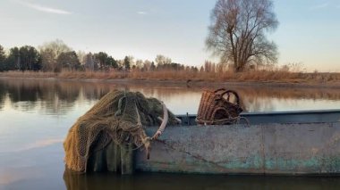 Nehir kıyısında eski bir balıkçı teknesi dağınık olta takımı, yıpranmış bir ağ ile kaplı ve sakin su ve doğa manzarası karşı hasır sepet