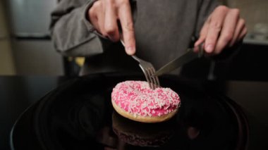 Tatlı gurmelerinin komik durumları. Elleri yakın plan, çatal ve bıçak kullanarak siyah tabakta beyaz parçacıklar serpiştirilmiş pembe bir donut kesiyorlar..