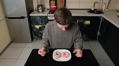 Genç bir adam masada oturuyor, iki donut kokluyor ve inceliyor, hangisini yiyeceğini seçmekte zorlanıyor, hoşgörü ve yemek arzusunu temsil ediyor.