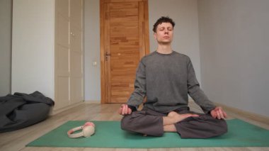 Evdeki yaşam alanında yerde oturup meditasyon yapan bir adam. Nilüfer pozisyonunda oturan sakin genç erkek yoga yapıyor.