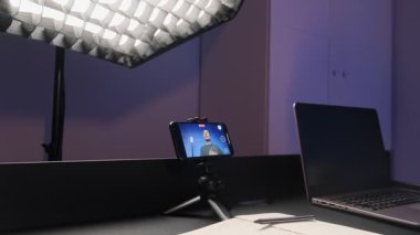 Erkek blogger video içeriğini profesyonel stüdyo ışığı altında akıllı telefonla kaydediyor, dizüstü bilgisayarla masaya oturuyor, ev ortamında modern dijital içerik oluşturulmasını sergiliyor.