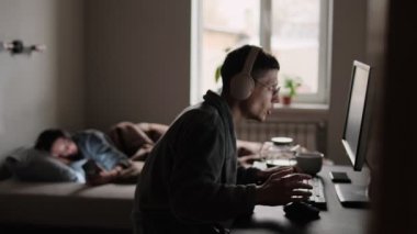 Bilgisayarda kulaklık ve gözlüklerle duygusal oyun oynayan bir adam, yatakta telefonu kaydıran bir kadın, her ikisi de ekrana kapılmış, doğal ışıkta yakalanmış, modern çift yaşamındaki kopukluğu yansıtan.