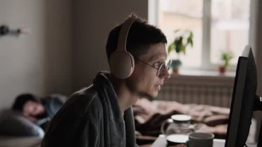 Bilgisayarda kulaklık ve gözlüklerle duygusal oyun oynayan bir adam, yatakta telefonu kaydıran bir kadın, her ikisi de ekrana kapılmış, doğal ışıkta yakalanmış, modern çift yaşamındaki kopukluğu yansıtan.