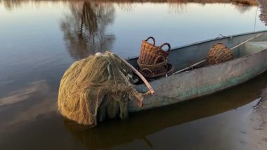 Nehir kıyısına yanaşmış eski bir balıkçı teknesi, ağlar, hasır sepet ve balıkçı teçhizatı ile dolu, sakin ve kırsal bir atmosfer yaratıyor.