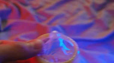 El ele tutuşan prezervatif yakın plan. Prezervatifli Güvenli Seks Konsepti. Arka planda, yatak renkli mahrem akşam ışığı ile aydınlatılır.