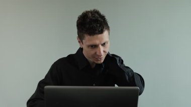 Dizüstü bilgisayarda çalışırken boynu ağrıyan bir adam. Rahatsiz çalisma pozisyonu boyun incinmesine neden oluyor. Gri arka planda siyah gömlekli bir ofis çalışanı.