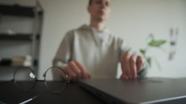 Genç serbest çalışan, evden çalışmaya başlar, dizüstü bilgisayarı açar, gözlük takar ve uzaktan kumandalı işe başlar, rahat bir iç mekanda, dijital çalışma alanına ve modern ofis rutinine odaklanır.