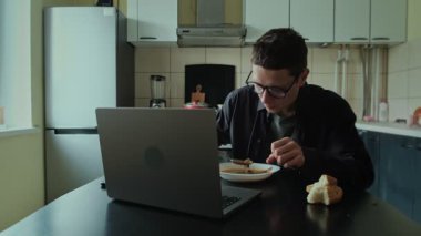 Koyu renk tişörtlü bir adam mutfakta dizüstü bilgisayarla çalışırken kahvaltı yapıyor, ev içi yemeklerle uzaktan kumanda yapıyor.