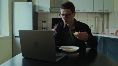 Genç adam bir kase çorbayla masada oturuyor, yemek yemek ve ev mutfağında dizüstü bilgisayar üzerinde çalışmak arasında gidip geliyor. Ciddi şekilde konsantre olmuş erkek serbest yemek yiyor, teslim tarihleri yanıyor.