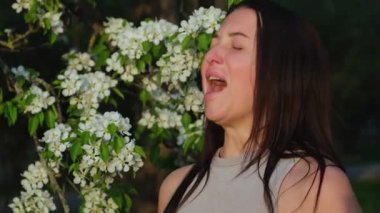 Sezonluk Alerji nedeniyle Hapşıran Genç Kadın. Esmer, Çiçek açan ağacın yanında duruyor ve Burun akıntısından muzdarip. Polene karşı alerjik reaksiyon. Beyaz peçete kullanan hasta beyaz kadın.