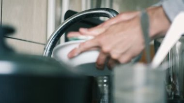 Ev mutfağında, sığ bir alan derinliği ve yumuşak ışıklandırmayla metal musluğun altında beyaz tabağı yıkayan ellerin yakın çekimi.