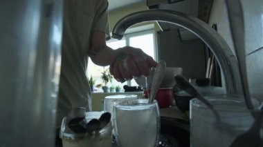 Bulaşık deterjanıyla sünger yakın plan. Mutfakta bulaşıklarla çevrili kirli tabakları temizlemeye başlıyor. Gerçekçi, dağınık ev rutini sahnesi.