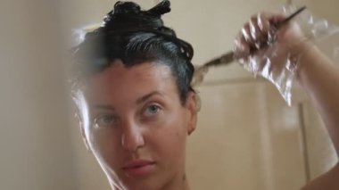Genç bir kadın banyodaki aynaya odaklanmış bir ifadeyle bakarken saçını fırça ve eldiven kullanarak boyuyor.
