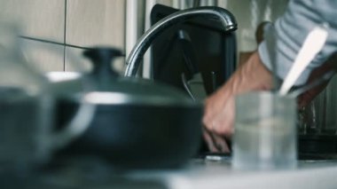 Bulaşık deterjanıyla sünger yakın plan. Mutfakta bulaşıklarla çevrili kirli tabakları temizlemeye başlıyor. Gerçekçi, dağınık ev rutini sahnesi.