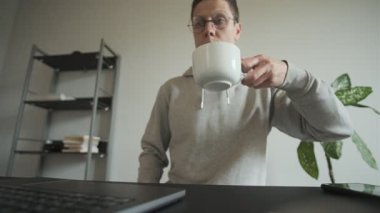 İçişleri Bakanlığı 'ndaki bir bardaktan kahvesini yudumlayan adam bilgisayarlı bir masanın arkasında. Freelancer rutini. Gün ışığı, ev içi..