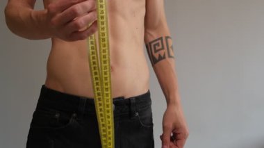 Bir adamın belini mezurayla ölçerken, sayıya memnuniyetle bakarken ve sonuçtan memnun olduğunu göstermek için baş parmak hareketinde bulunurken. Kilo verme süreci, fitness başarısı veya sağlıklı yaşam tarzı takibi kavramı.