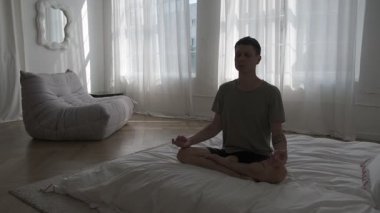 Büyük pencereleri ve doğal ışığı olan parlak minimalist bir dairede stresi azaltmak için meditasyon yapan bir adam.