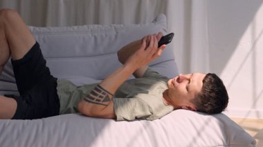 Genç bir adam modern bir evde, bir kanepede uzanıyor, akıllı telefonuyla kayıyor. Ekran bağımlılığı, sosyal medya bağımlılığı ve günlük hayatta sonsuz kısa video tüketimi kavramı.