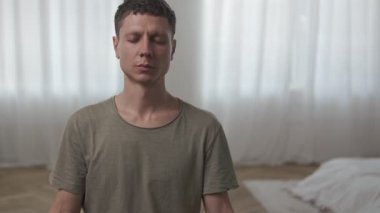 Genç adam modern koltukta gözleri kapalı meditasyon yapıyor. Minimalist iç mekanda iç huzuru buluyor..