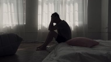 Panik ve zihinsel baskı altında, karanlık odada yatakta oturan endişeli bir adamın silueti.