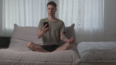 Genç adam meditasyonu akıllı telefondan öğreniyor ve pratik yapmaya başlıyor. Dikkatli rutin, çevrimiçi ruhsal kurs, modern zihinsel sağlık alışkanlıkları
