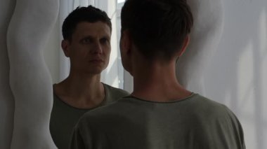 Genç adam parmaklarıyla dudaklarını çekerek aynaya gülümsemeye çalışıyor. Depresyon, zihinsel sağlık sorunları ve duygusal acı kavramı. Yumuşak ışıkla minimalist iç mekan.