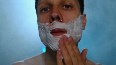 Genç adam düşük ışıkta yüzüne tıraş kremi sürüyor mavi arka plan ve yumuşak dumanla. Cilt bakımı ve bakım rutini.