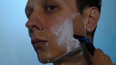 Genç adam tıraş kremi sürdükten sonra yüzünü jiletle tıraş etmeye başlar. Mavi arka plan ve sisli karamsar ışıkta yakın çekim. Samimi tımar anı.