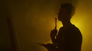 Smoky Stüdyo 'daki düşünceli sanatçının silueti tuvalin önünde duruyor, ilk fırça darbesinden önce tereddüt ediyor..