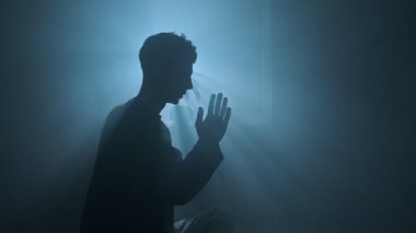 Mavi sisli odada dua eden bir adam ilahi ışık huzmeleriyle pencereden süzülen, ruhani bir siluet sahnesi