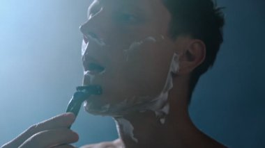 Genç adam düşük ışıkta yüzüne tıraş kremi sürüyor mavi arka plan ve yumuşak dumanla. Cilt bakımı ve bakım rutini.