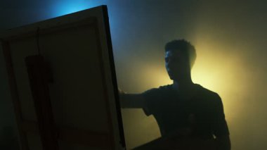 Sinema sehpasında resim yapan genç bir erkek sanatçı. Huysuz sarı ve mavi arkaplan, yan görüş.