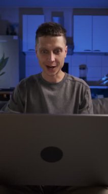 Adam dizüstü bilgisayarından iyi haberler alıyor, kutlama yapıyor ve gülümsüyor, gündelik giysiler, ev mutfağında koltukta oturuyor, gerçek hayat anı.