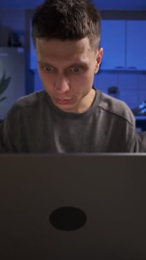 Adam dizüstü bilgisayarından iyi haberler alıyor, kutlama yapıyor ve gülümsüyor, gündelik giysiler, ev mutfağında koltukta oturuyor, gerçek hayat anı.