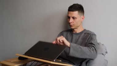 Gri kapüşonlu genç adam gri duvarlı yatakta oturuyor ve işe başlamak için bir dizüstü bilgisayar açıyor. Serbest çalışma, uzaktan çalışma, çevrimiçi işler, verimlilik ve ev yaşamından çalışma kavramı.