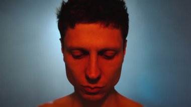 Kırmızı ışıkla aydınlatılan bir adam mavi dumanlı bir arkaplanda öylece durur, gözlerini indirir ve bir şeyi anlamaya çalışır gibi kameraya bakar..