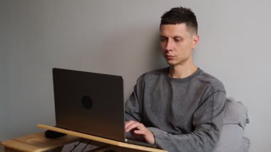 Gri kapüşonlu genç adam evde dizüstü bilgisayarla çalışıyor. Serbest çalışma, uzaktan çalışma, çevrimiçi işler, üretkenlik ve ev hayatı kavramı. Yakın çekim..