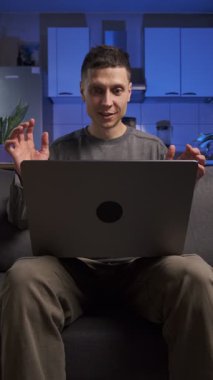 Adam dizüstü bilgisayarından iyi haberler alıyor, kutlama yapıyor ve gülümsüyor, gündelik giysiler, ev mutfağında koltukta oturuyor, gerçek hayat anı.