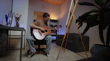 Yaratıcı otantik ev dekorasyonundaki genç adam akustik gitar öğrenme müziği çalıyor. Resimli tuvali ve arka planda masası olan rahat bir yaşam tarzı..