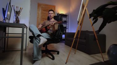 Yaratıcı otantik ev dekorasyonundaki genç adam akustik gitar öğrenme müziği çalıyor. Resimli tuvali ve arka planda masası olan rahat bir yaşam tarzı..