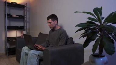 Genç serbest çalışan, kötü duruş nedeniyle boyun ağrısı çekiyor. Kanepede kambur oturuyor ve otantik ev ortamında dizüstü bilgisayarla çalışıyor. Yerleşik işlerin, aygıtların rahatsızlığının ve ev ofis yaşam tarzının vurgulanması.