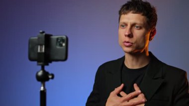 Takım elbiseli genç adam RGB renkli arka planlı akıllı telefon tripodunda motivasyon videosu kaydediyor..