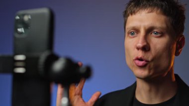 Takım elbiseli genç adam RGB renkli arka planlı akıllı telefon tripodunda motivasyon videosu kaydediyor..