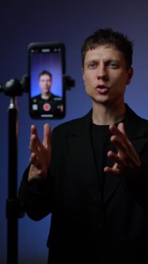 Takım elbiseli genç adam RGB renkli arka planlı akıllı telefon tripodunda motivasyon videosu kaydediyor..