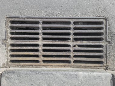 Asfalt kaldırıma gömülü dikdörtgen metal rögar ızgarası..