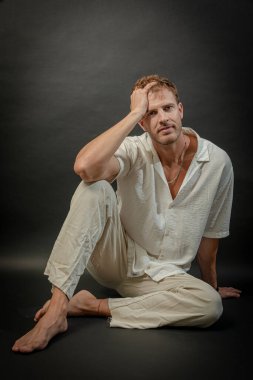 Göz alıcı, mavi gözlü, zarif bir erkek model, siyah bir arka plana karşı zarif bir poz veriyor. İnce bir keten gömlek ve nötr renkli kumaş pantolon giyiyor, sofistike ve zamansız bir tarz sergiliyor..